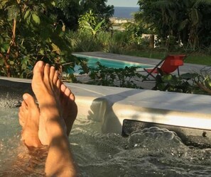 A person's feet in a hot tub with a pool and greenery in the background. - Poivre & Citronnelle (Saint-André)