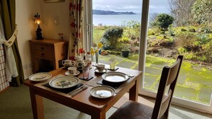 Mesa de jantar posta com pratos, chávenas e uma lâmpada, com vista para um jardim e para o mar.