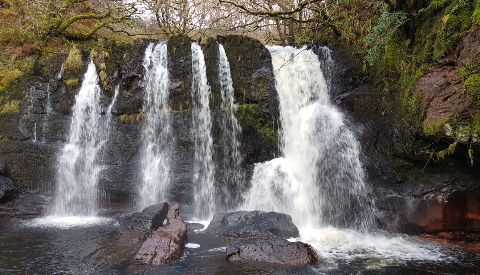 A waterfall with multiple streams cascading over rocks, surrounded by mossy vegetation.