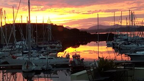 Uma marina com barcos atracados, águas calmas refletindo o pôr do sol, e montanhas ao fundo.