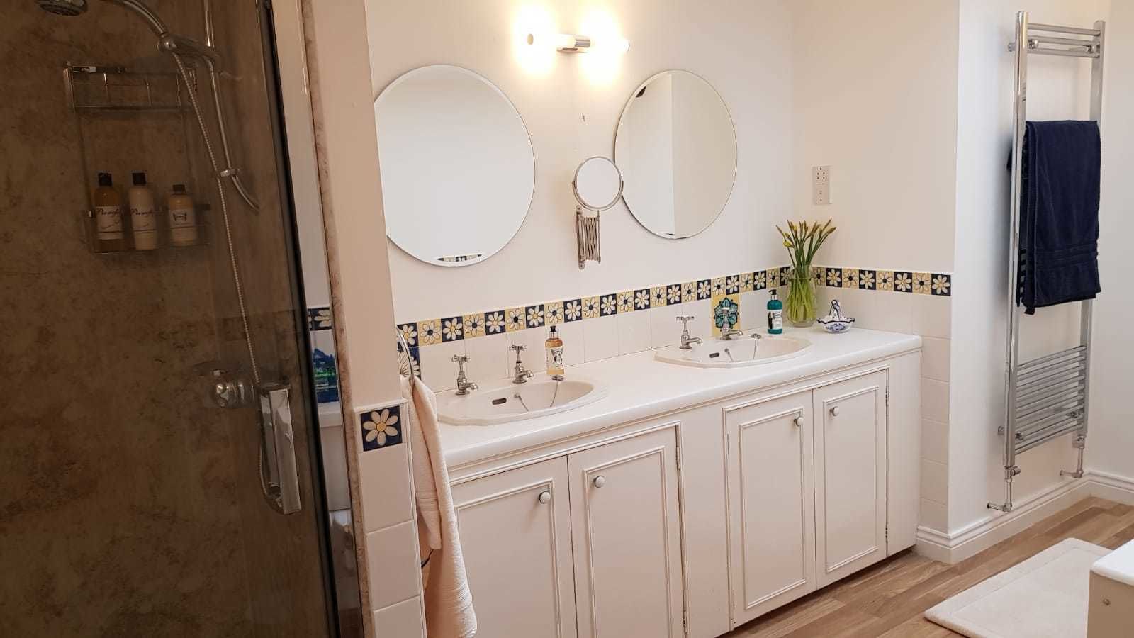 Casa de banho com lavatório duplo, duas espelhas, chuveiro e porta-toalhas.