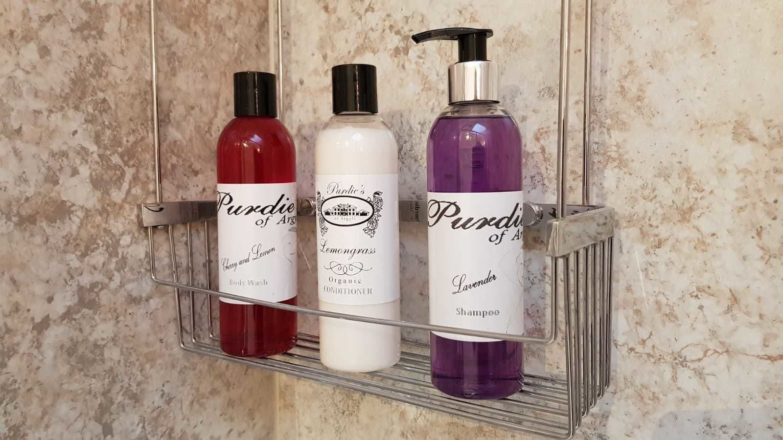 A bathroom shelf holding three bottles of Purdie of Argyl body wash, organic conditioner, and lavender shampoo.