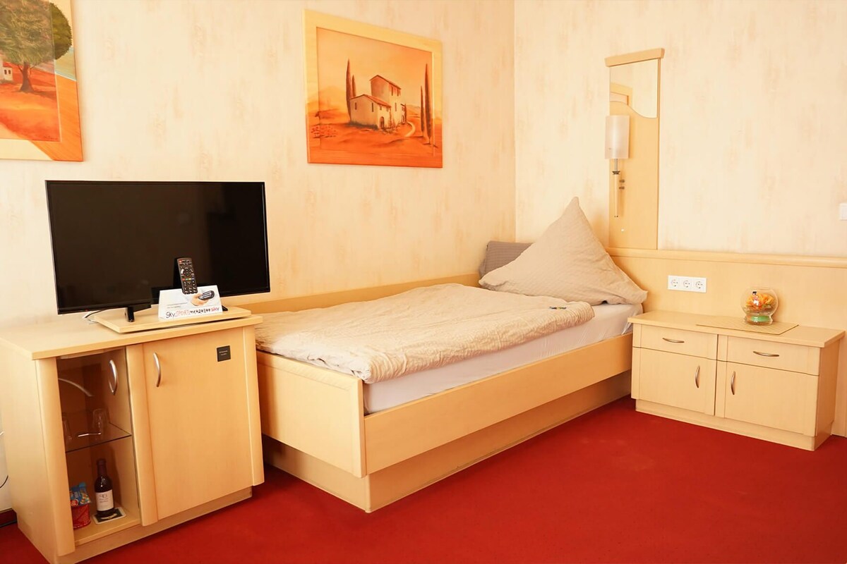 A single-bed room with a TV, a small cabinet with wine, a mirror, and artwork on the walls.
