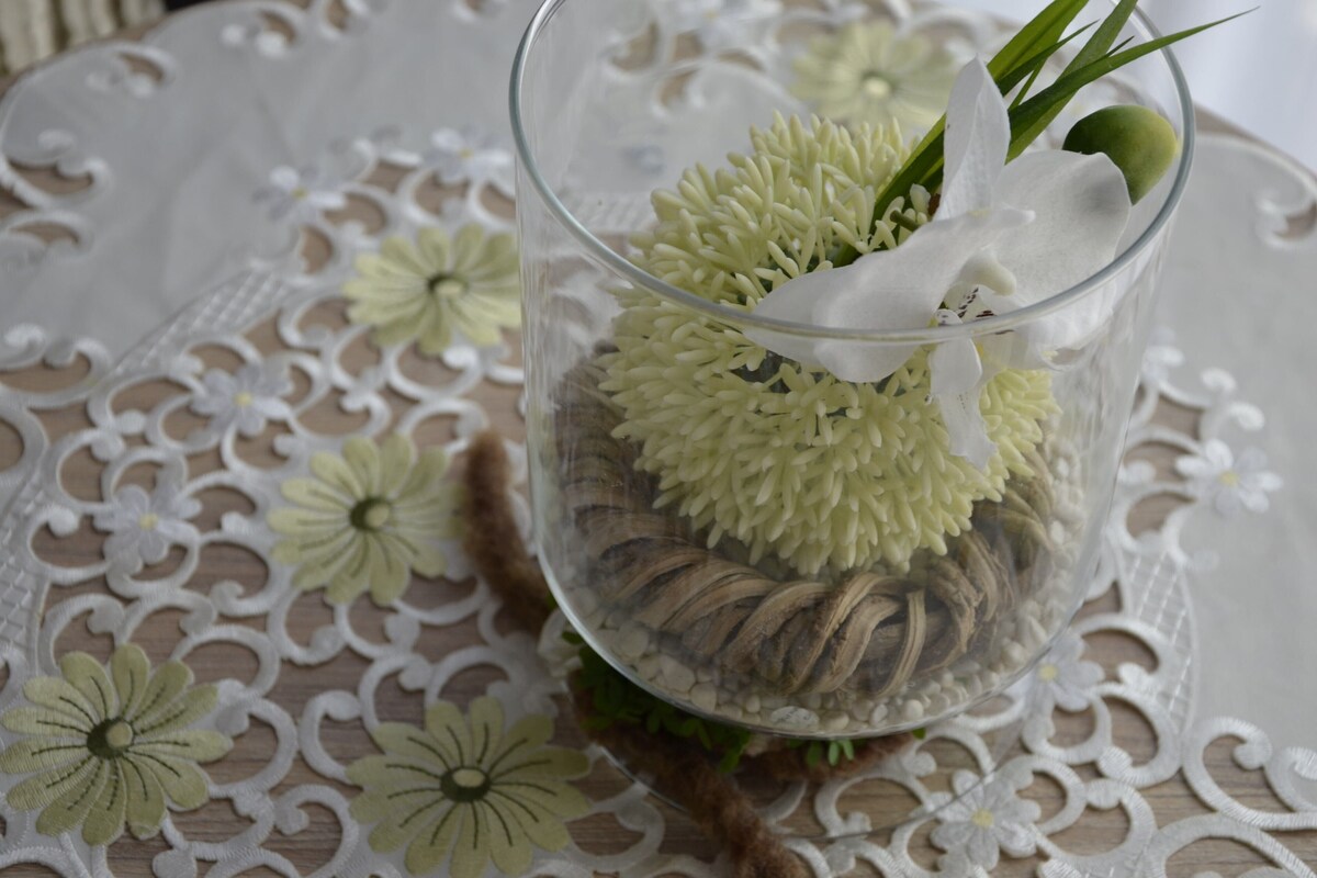A clear glass vase with dried flowers and green foliage on a lace tablecloth.