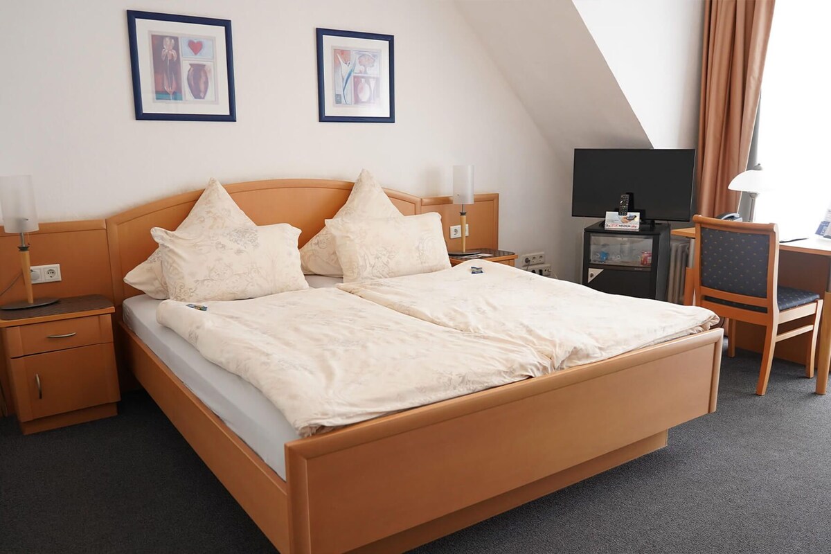 A hotel room with a bed, bedside tables, a desk, a chair, and two framed pictures on the wall.