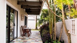 Eine überdachte Terrasse mit Schachbrettmuster, einem Tisch mit Stühlen und Blick auf einen Garten mit verschiedenen Pflanzen und Bäumen.