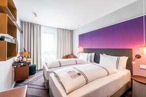 Een hotelkamer met twee bedden, een bureau, een stoel, een lamp en een groot raam met gordijnen.