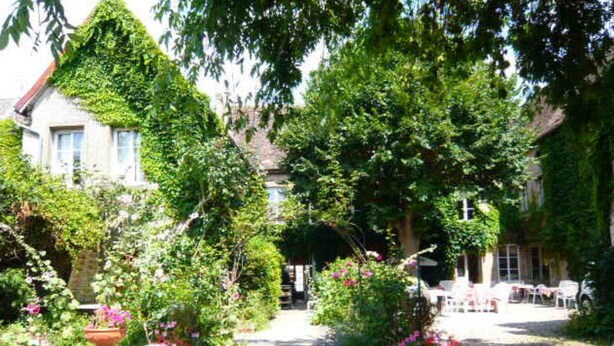 En sti gennem en have fører til en bygning i traditionel arkitektonisk stil, omgivet af frodig vegetation og blomstrende planter.