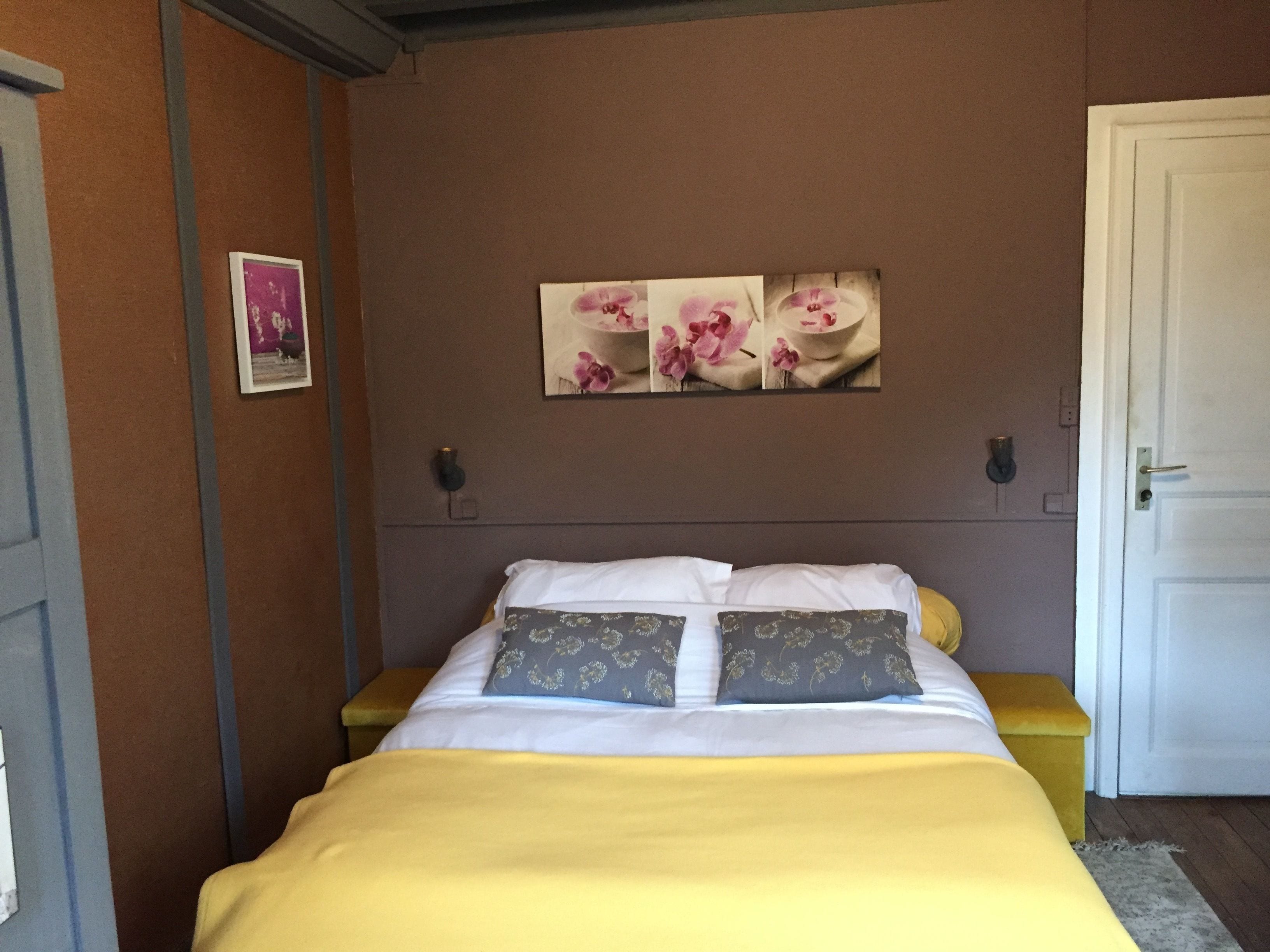 Ein Schlafzimmer mit einem Bett, einer gelben Decke, zwei Kissen mit Blumenmustern und einem Wandbild von rosa Blumen.