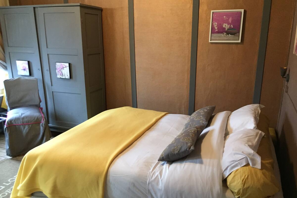 A neatly made bed with a yellow blanket and pillows, a gray wardrobe, and a framed picture on the wall.