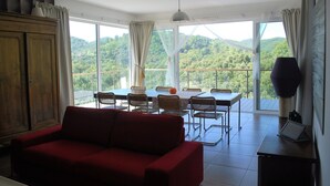A room with a red sofa, dining table with chairs, and large windows offering a view of green hills.