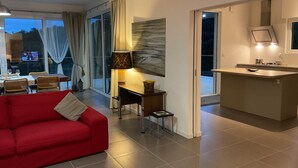 A modern living room with a red sofa, a dining area with a table and chairs, a kitchen with a counter and sink, and a large window with curtains.