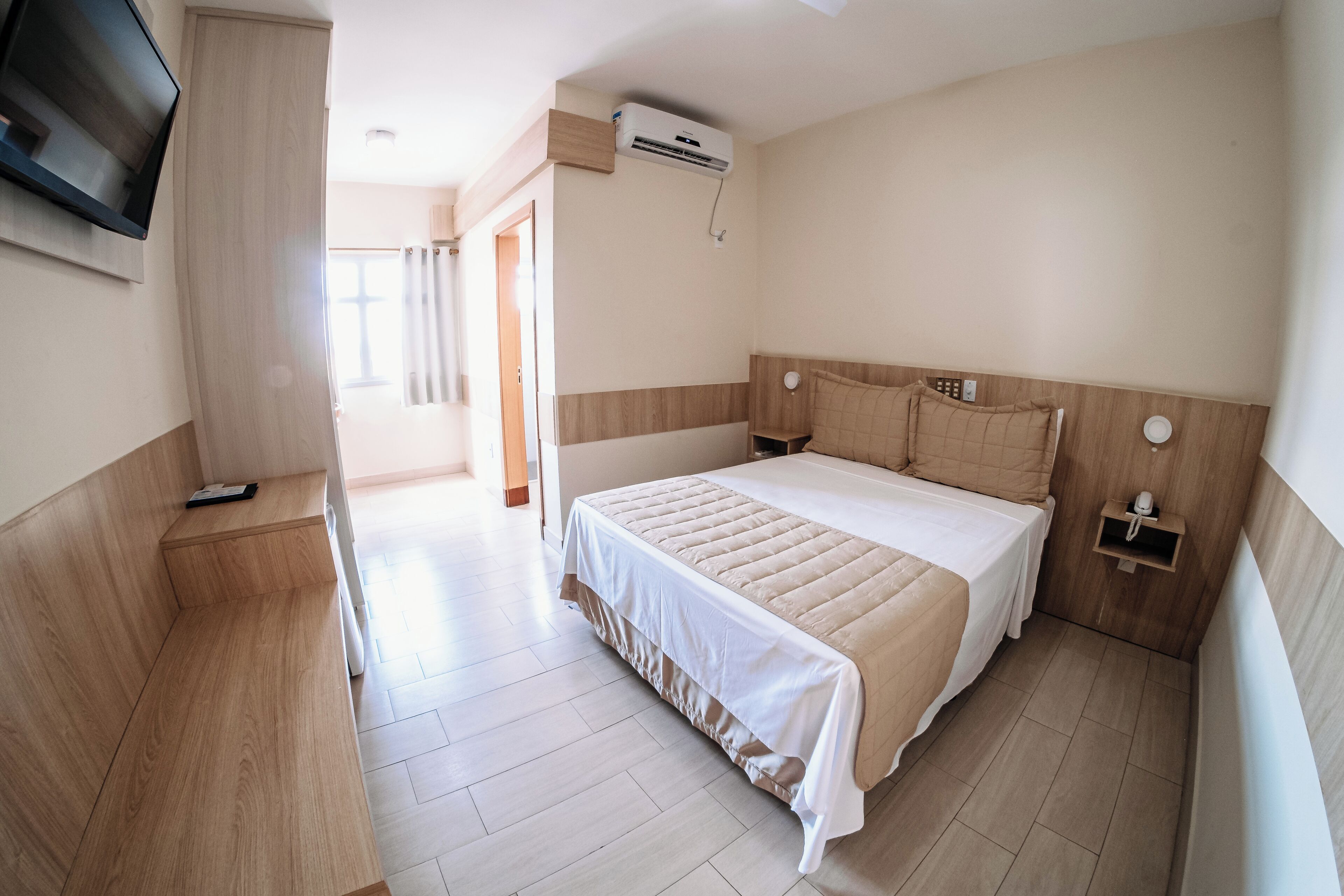 A hotel room with a bed, bedside tables, a TV mounted on the wall, and an air conditioning unit.