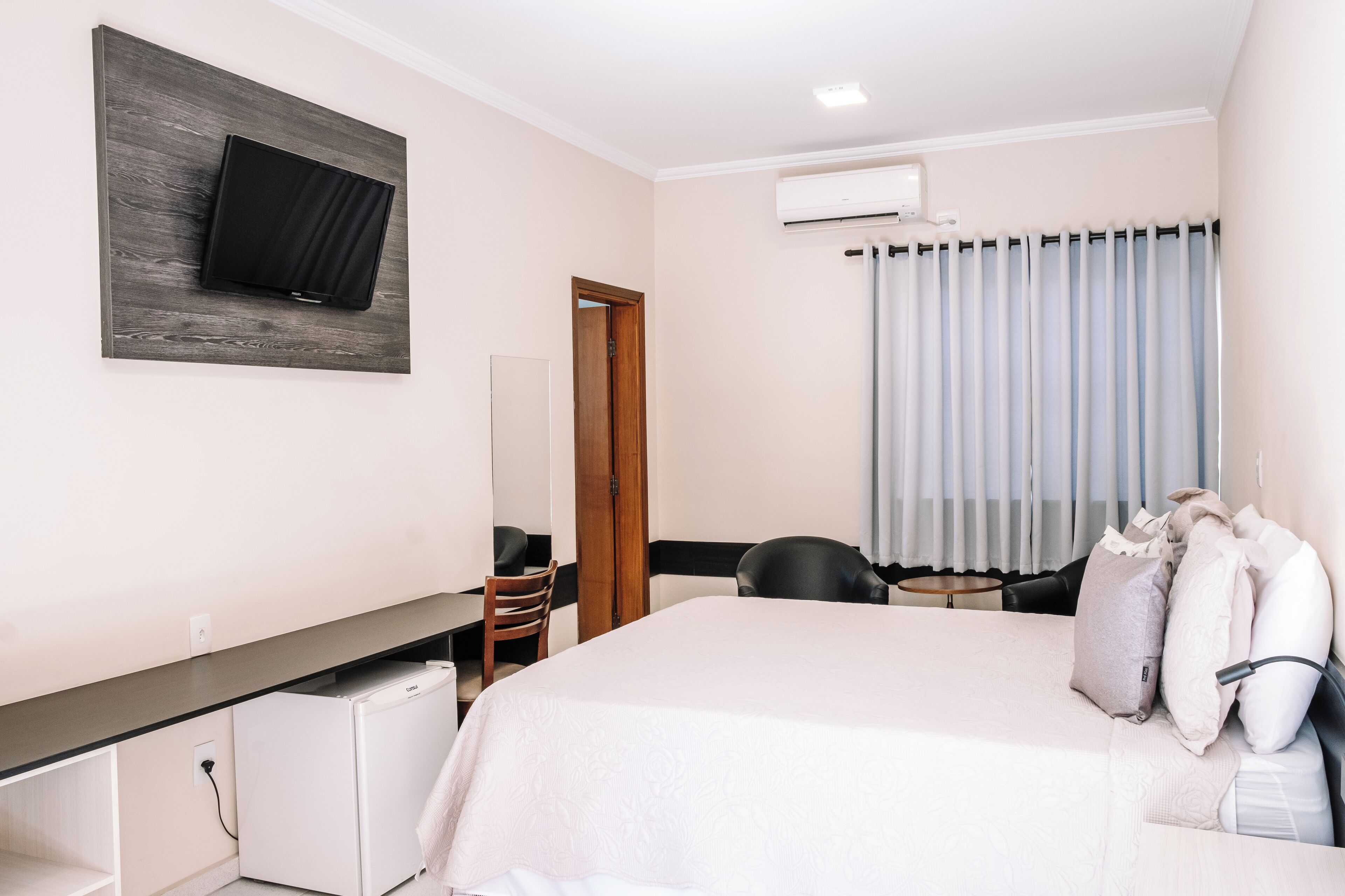 Ein Hotelzimmer mit einem Bett, einem Schreibtisch, einem Stuhl, einem an der Wand befestigten Fernseher und einem Fenster mit Vorhängen.