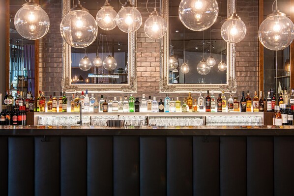 Un bar con varias botellas de licor y vasos, lámparas esféricas colgantes y un espejo grande.