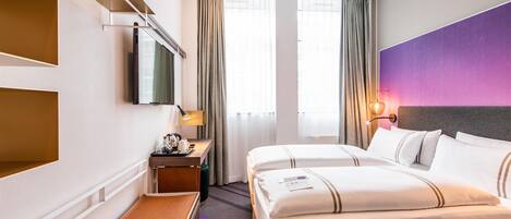 Habitación de hotel con cama, escritorio con lámpara, televisor y ventana con cortinas.