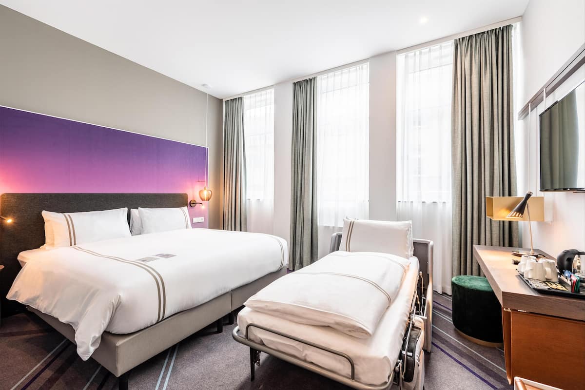 Ein Hotelzimmer mit zwei Betten, einem Schreibtisch, einem Stuhl, einem Fernseher und einem großen Fenster mit Vorhängen.