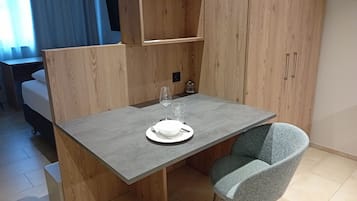 A compact room with a wooden wardrobe, a small dining table with a chair, and a bed in the background.