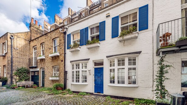 Une rangée de maisons blanches mitoyennes aux volets bleus, une rue pavée et un banc au premier plan.