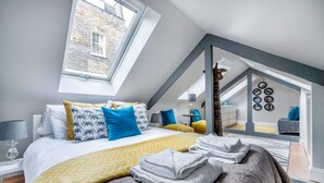 Une chambre moderne avec un puits de lumière, un lit agrémenté d’oreillers à motifs, un plaid jaune et une vue sur le paysage urbain par la fenêtre.