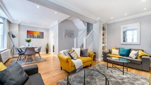 Un vaste espace de vie comprenant un canapé jaune, un canapé gris et une table basse en verre. Un escalier mène à un autre niveau, et un coin repas est aménagé avec une table et des chaises.