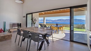 A modern dining area with a wooden table and chairs, a TV, and a view of the sea and mountains through large glass doors. - Résidence La Plage (Olmeto)