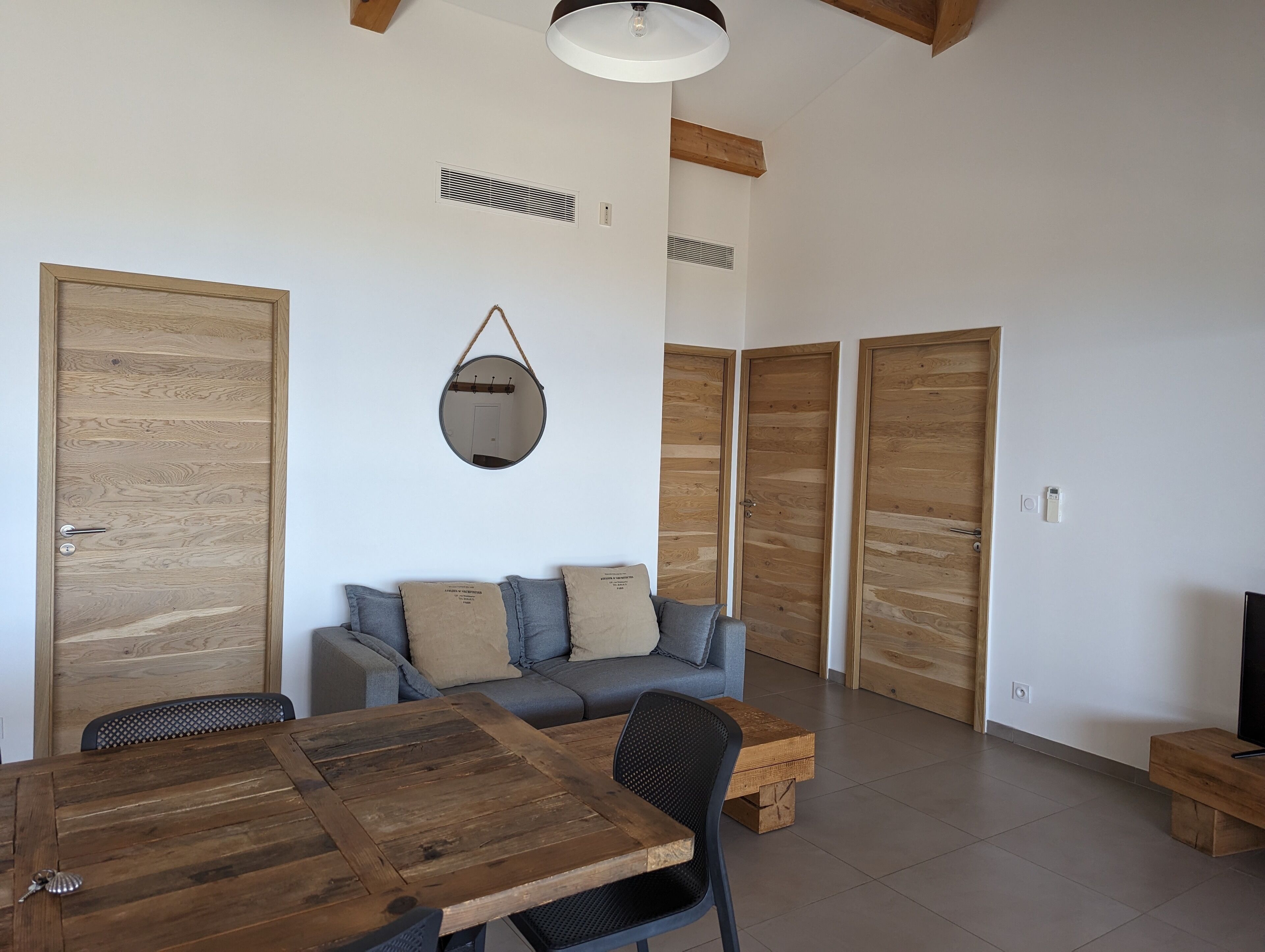 A modern living room with a wooden dining table, a sofa with cushions, and a hanging mirror.