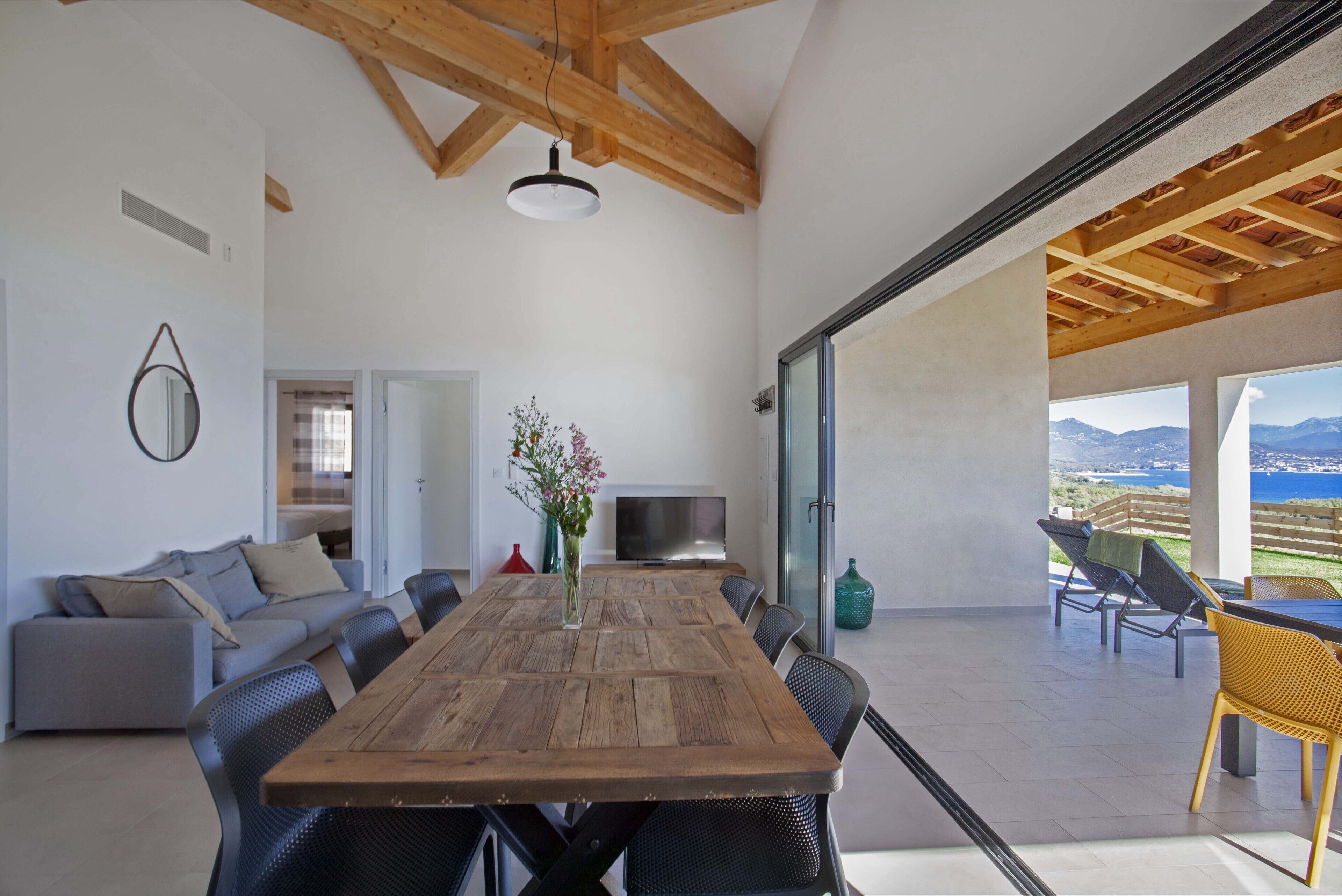 Uma sala de estar ampla com uma mesa de jantar de madeira, assentos confortáveis, uma televisão e vista para o exterior.