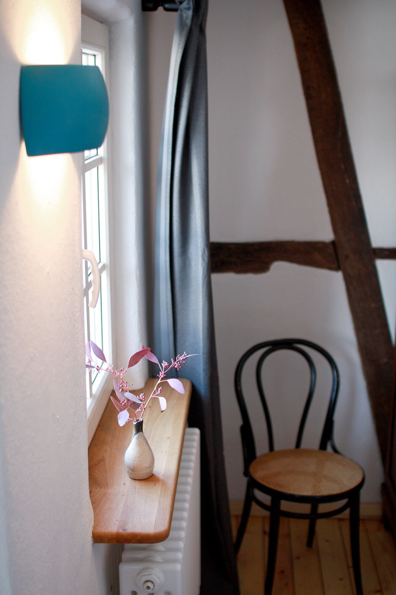 Una stanza con un tavolo in legno, un vaso con fiori rosa, una sedia nera, un termosifone e una finestra con tende.