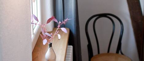 Una stanza con un tavolo in legno, un vaso con fiori rosa, una sedia nera, un termosifone e una finestra con tende.