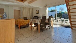 A spacious living area with a dining table, wicker chairs, and a sofa with a yellow blanket. There is a large window with curtains, a wooden ceiling, and a view of the outside. - Spacious villa near beach, 5 people, private garden (La Faute-sur-Mer)