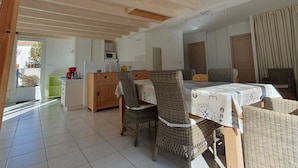A spacious dining area with a high ceiling, a kitchen in the background, and a view of the outdoors. - Spacious villa near beach, 5 people, private garden (La Faute-sur-Mer)