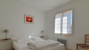 A bedroom with a bed, a chair, a radiator, a window, and a wall-mounted lamp. - Spacious villa near beach, 5 people, private garden (La Faute-sur-Mer)