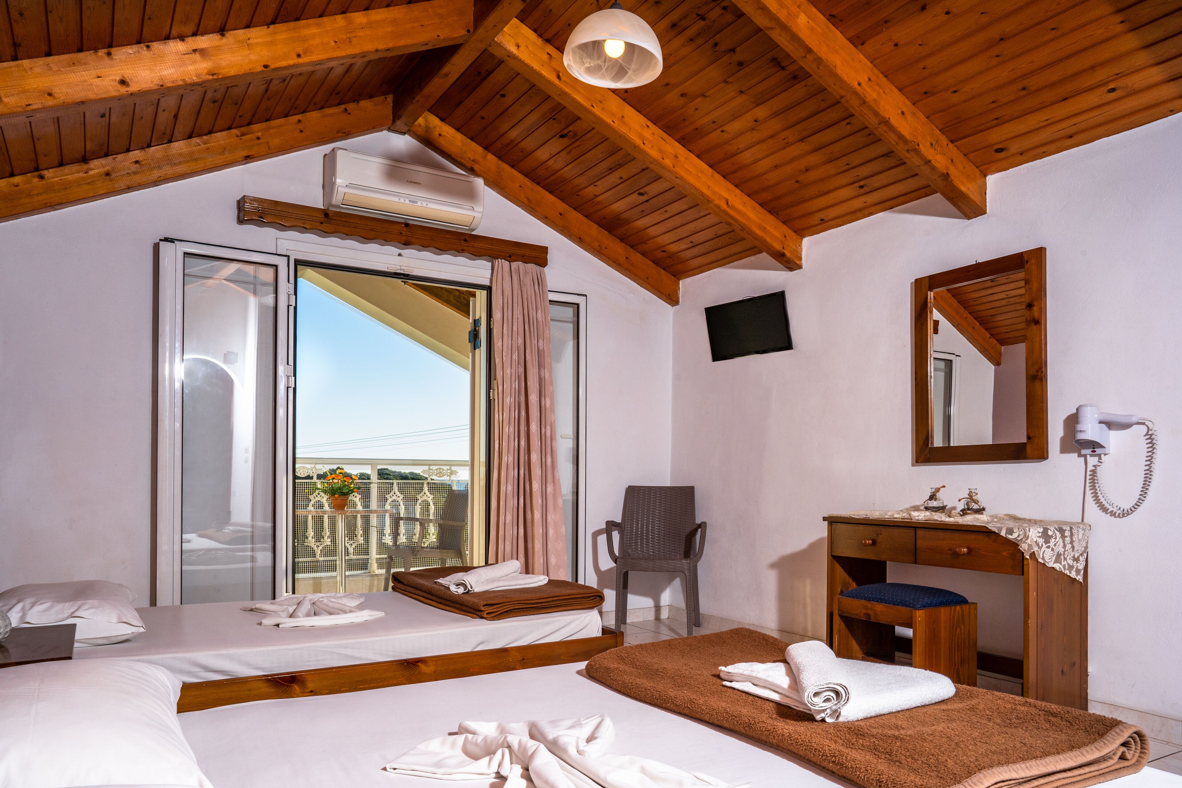 Une chambre d’hôtel avec deux lits, un plafond en bois, un balcon avec vue, un bureau avec un miroir et un sèche-cheveux.
