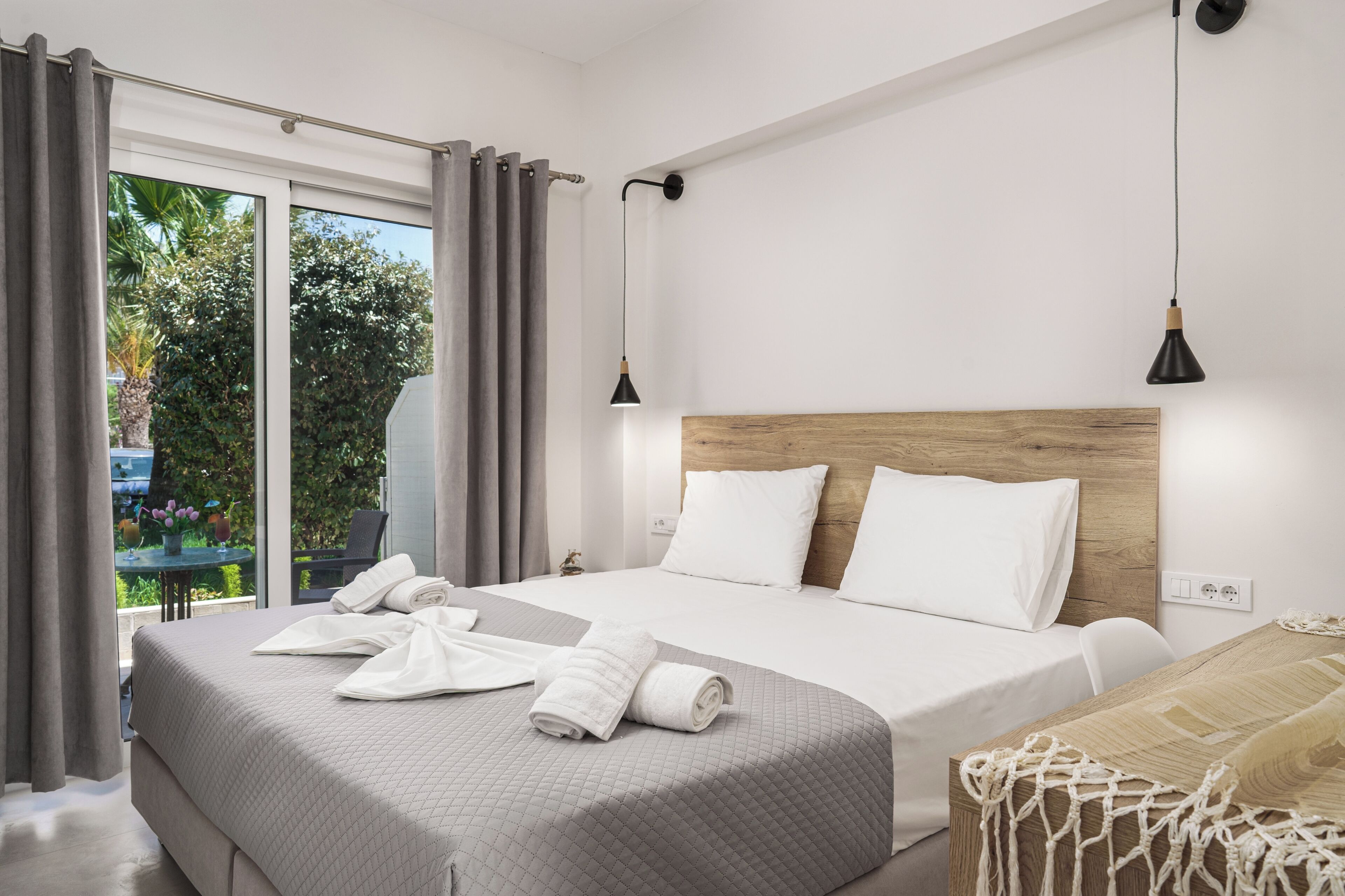 A modern hotel room with a large bed, a wooden headboard, and a view of greenery through the window.
