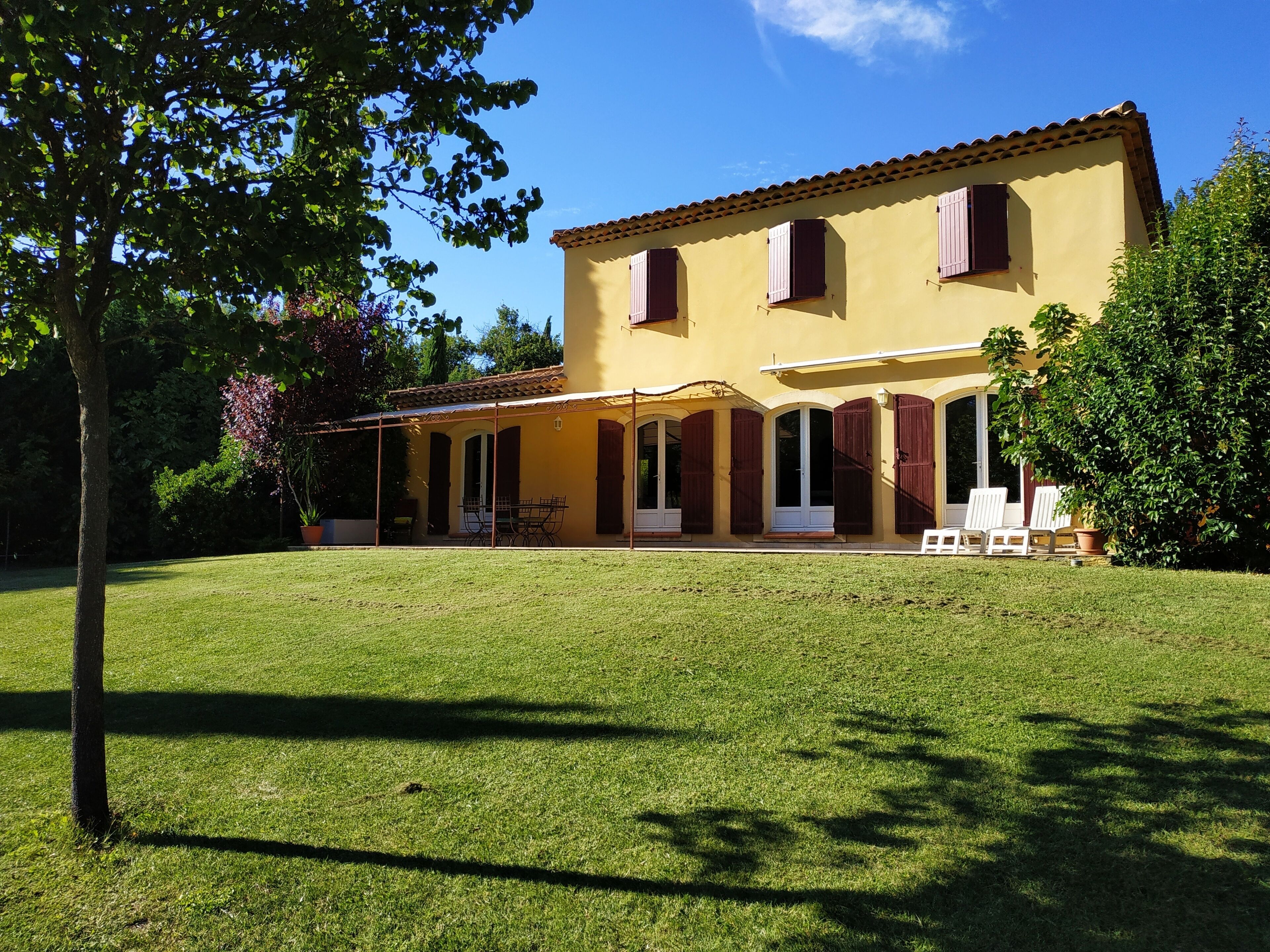 Un edifici de dues plantes amb façana groga, diverses finestres amb persianes i una zona de pati coberta amb seients exteriors.