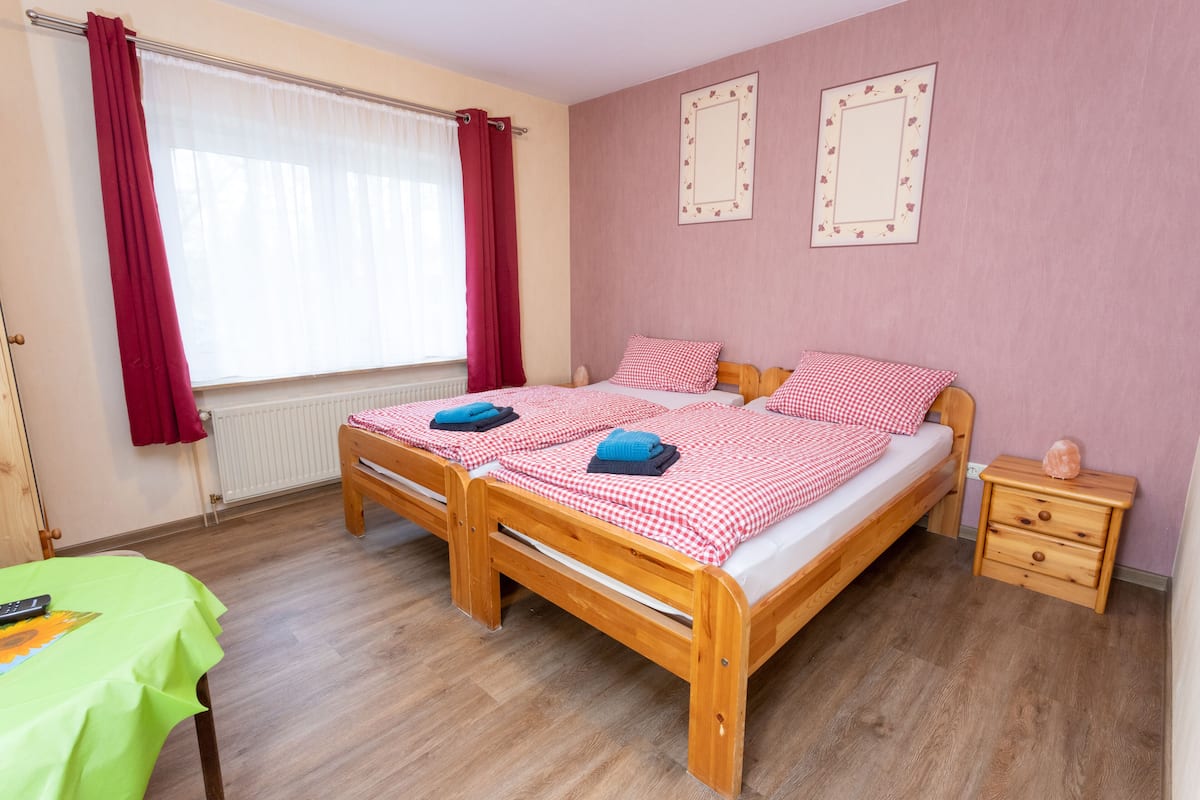 A double bed with checkered bedding, a wooden nightstand, a table with a green tablecloth, and a window with red curtains.