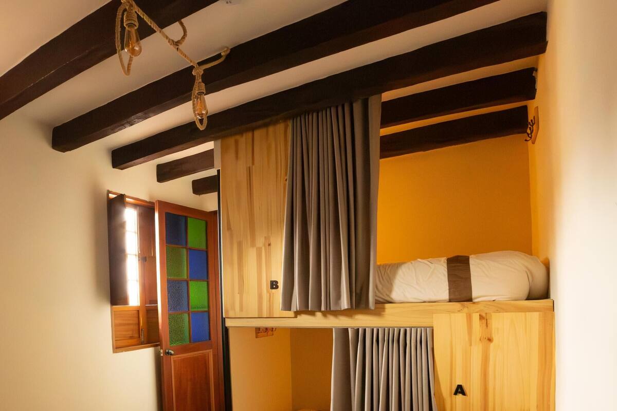 Habitació amb terra de fusta, un llit superposat amb cortines, un armari de fusta i una taula petita.