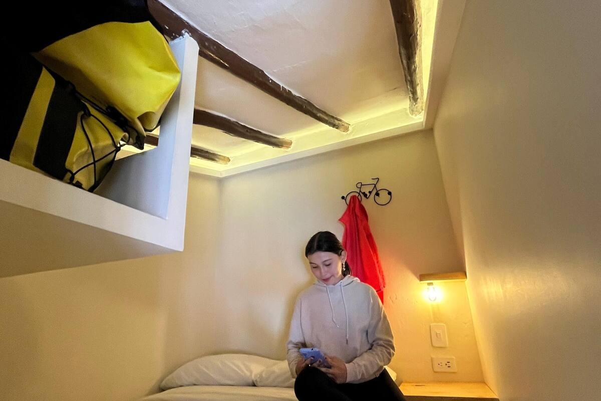 Una persona asseguda en un llit d'una habitació amb sostre inclinat, una bicicleta penjada a la paret i una cortina vermella.