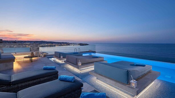 A rooftop infinity pool with lounge seating and a city view at dusk. - Seafront Apt Studio with Seaview, infinity pool, beachfront (Limenas Chersonisou)
