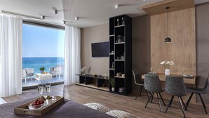A modern hotel room with a balcony overlooking the sea, a dining area with a table and chairs, a bookshelf, and a flat-screen TV mounted on the wall. - Seafront Apt Studio with Seaview, infinity pool, beachfront (Limenas Chersonisou)