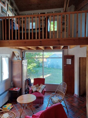A cozy room with a wooden ceiling, a red sofa, a wicker chair, and a view of a garden through a large window.