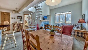 A spacious living area with a wooden dining table, chairs, a red sofa, a staircase, and a large window with a view of trees. - Luxury 2br + Loft, Lodge At Kirkwood Unbeatable Location 2 Bedroom Condo by RedAwning (Kirkwood)