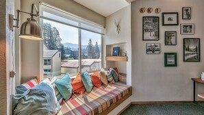 Een gezellige kamer met een ingebouwde bank, een raam met uitzicht op huizen en bomen, en een muur versierd met ingelijste foto’s en een woordmerk met ski-thema.