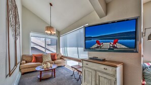 Een woonkamer met een grote televisie, een bank, een salontafel en uitzicht op een steiger met stoelen.