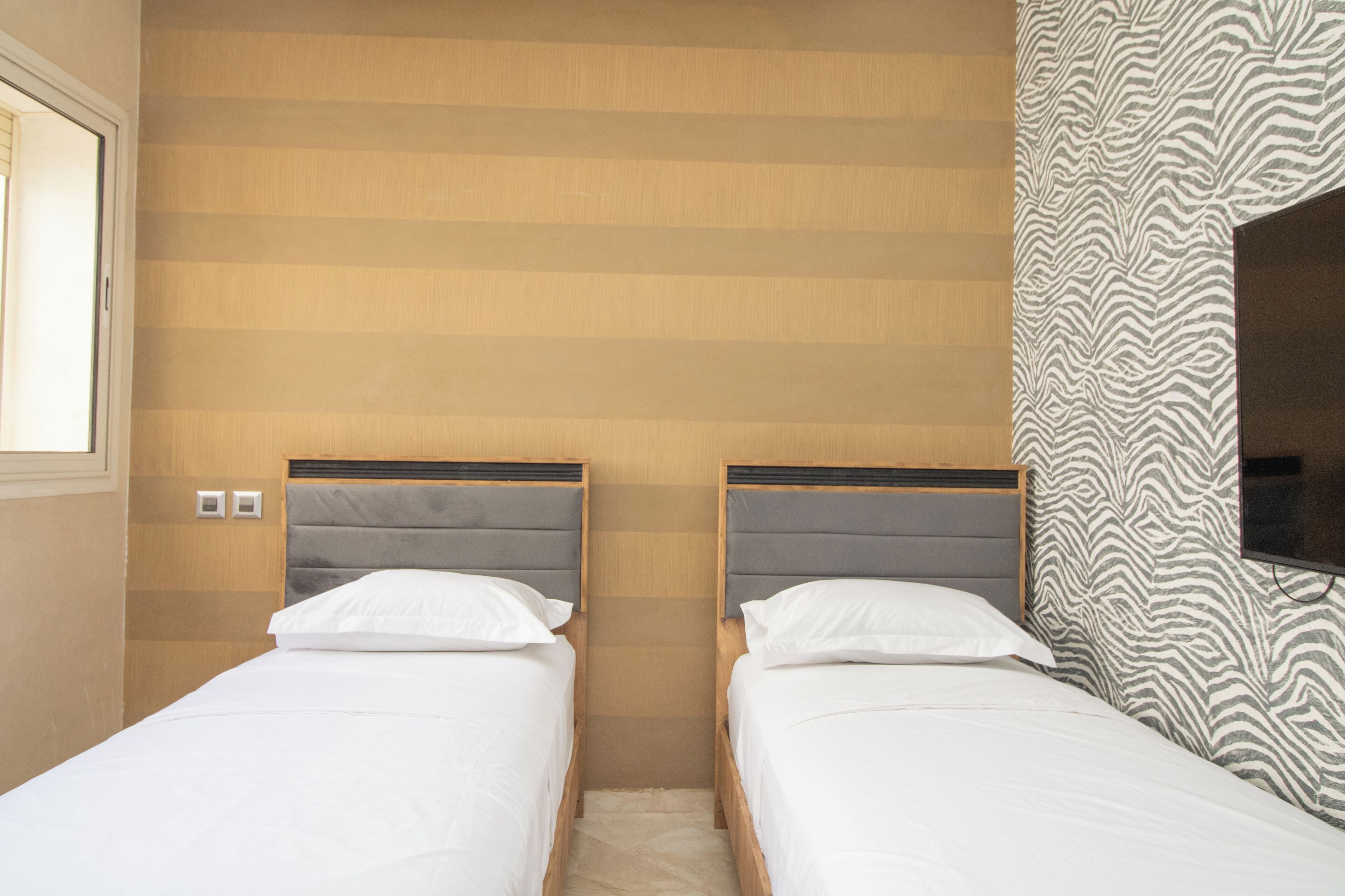 A hotel room with two single beds, wooden headboards, a patterned wall, and a television mounted on the wall.