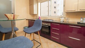 A modern kitchen with a glass dining table, two blue chairs, a stainless steel refrigerator, a gas stove, and maroon cabinets. - Studio de Luxe à Casablanca Aireport (Nouaceur)