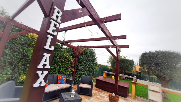 Een houten pergola met de tekst 'RELAX' erop, omringd door tuinmeubilair, waaronder een bank en een loungebank, voor een groene achtergrond.