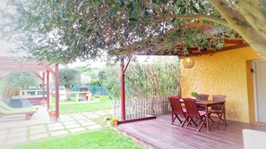 A covered patio with a wooden table and chairs, surrounded by a garden and a yellow wall.