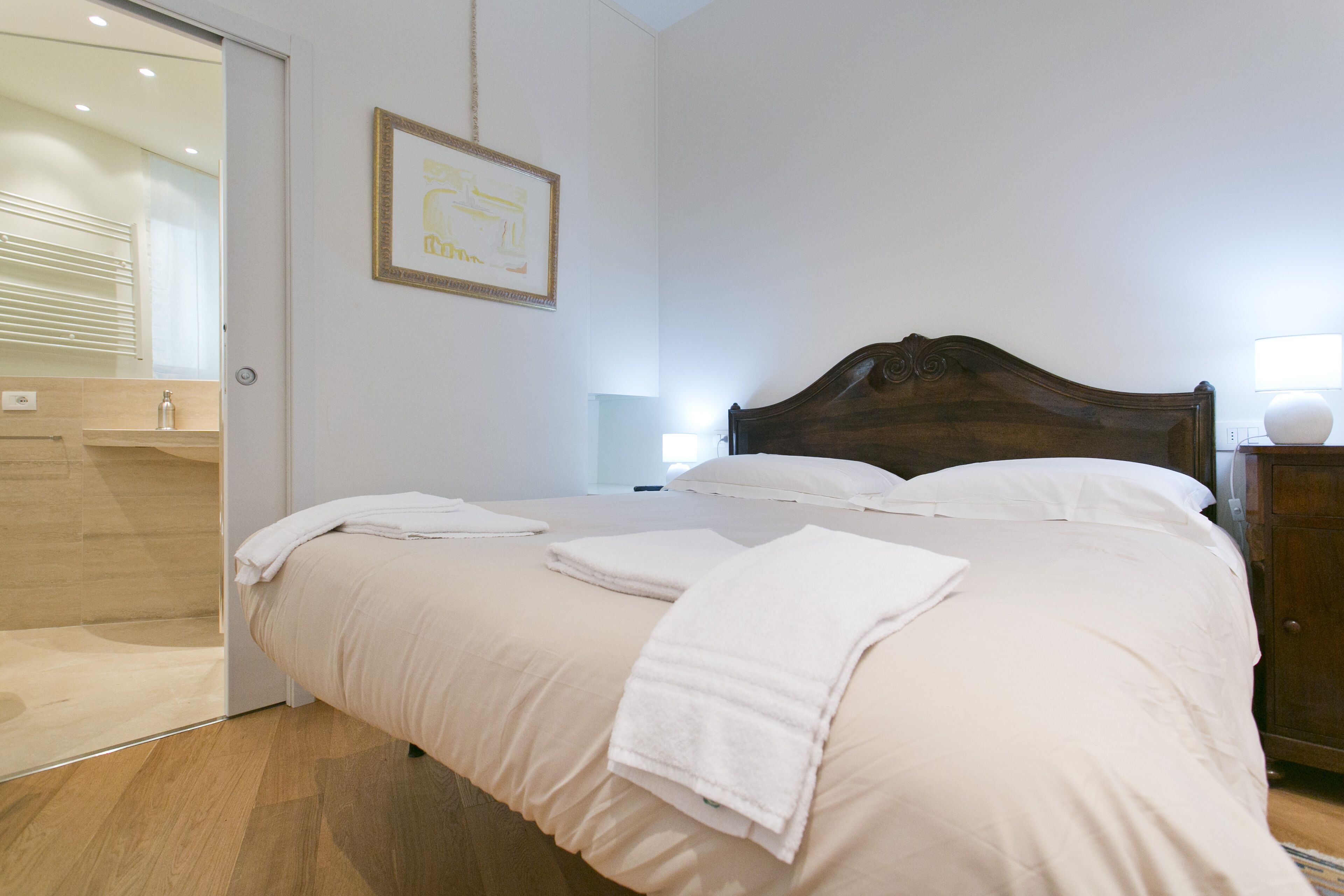 Cama bem arrumada com lençóis e toalhas brancas, cabeceira de madeira e mesa de cabeceira com abajur.